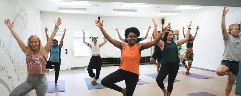 A full class at Soul Strong Yoga standing in tree pose, several students using blocks for support.